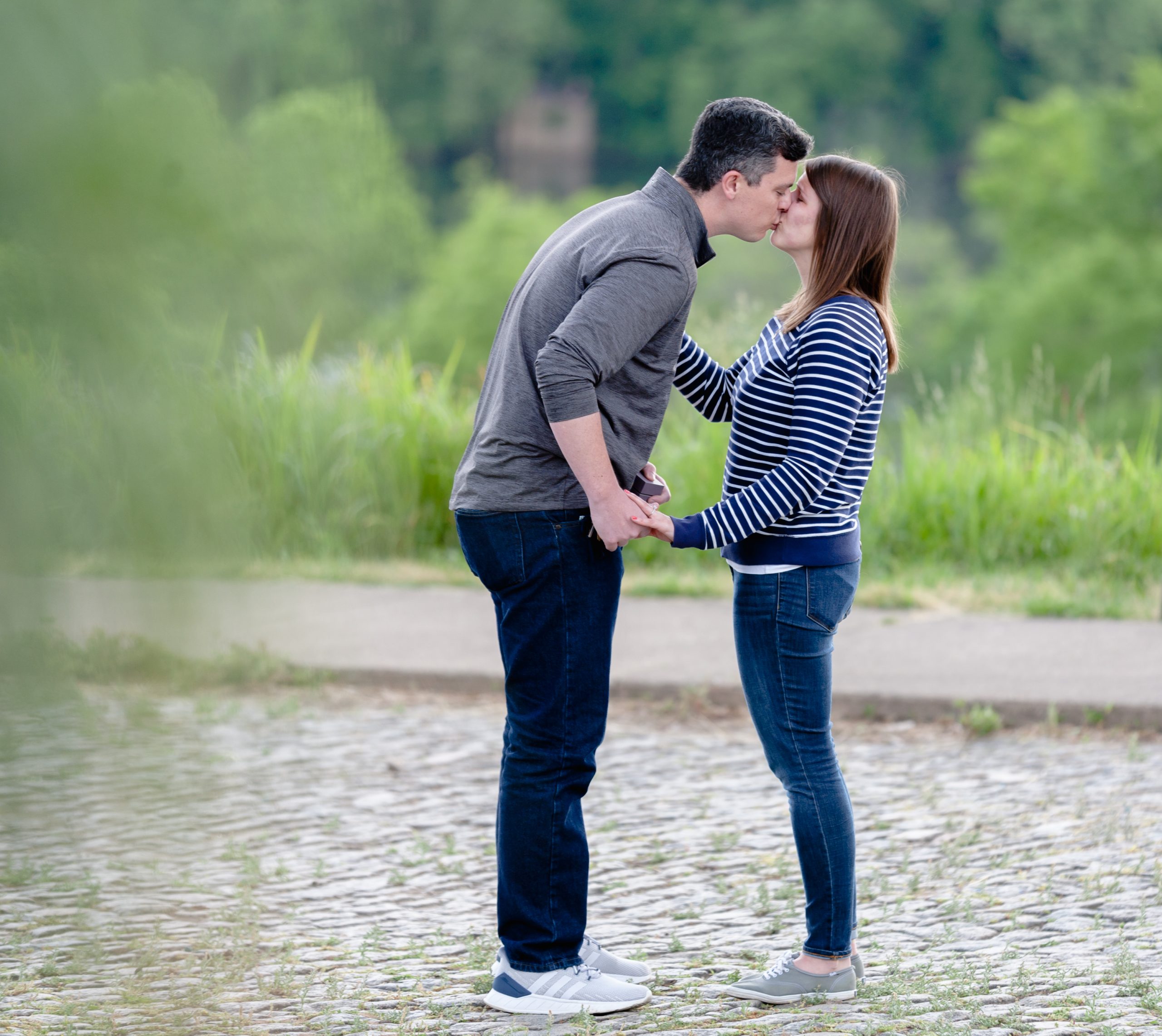Image of couple man and woman kissing during a wedding proposal caught on camera in the street of a local park with green urban forest behind